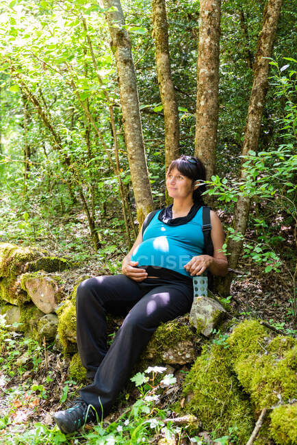 Pregnant female traveler sitting on mossy stones in green woods and having break during trekking in summer — Fotografia de Stock