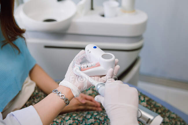 Crop unrecognizable female dental hygienist with prosthetics and electric toothbrush demonstrating patient how to brush teeth correctly during appointment in modern dental clinic — Foto stock