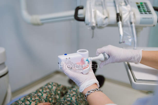 Crop unrecognizable female dental hygienist with prosthetics and electric toothbrush demonstrating patient how to brush teeth correctly during appointment in modern dental clinic - foto de stock