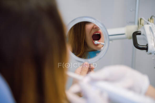 Урожай молодої жінки, яка сидить на стоматологічному стільці з відкритим ротом і дивиться на зуби в дзеркало під час відвідування професійного стоматолога в сучасній клініці — стокове фото