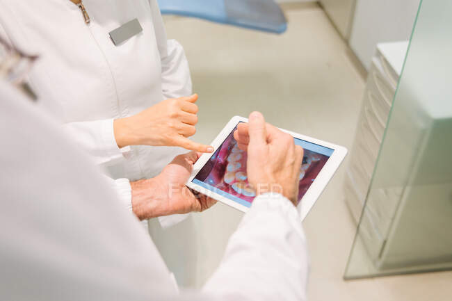 Dentistes anonymes en peignoirs médicaux examinant l'état des dents sur la radiographie sur tablette tout en travaillant ensemble dans une clinique dentaire moderne — Photo de stock