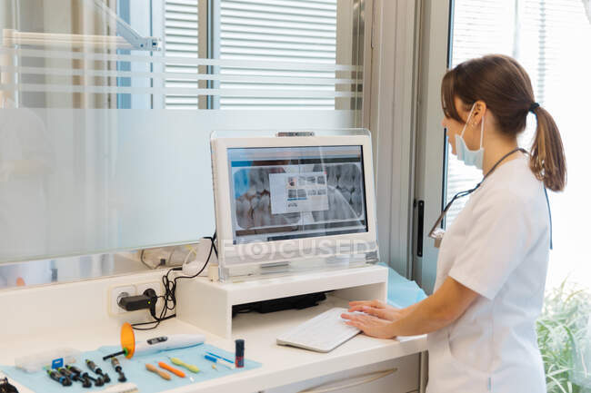 Side view competent female dentist in uniform examining x ray picture on screen in light modern dental clinic — Foto stock