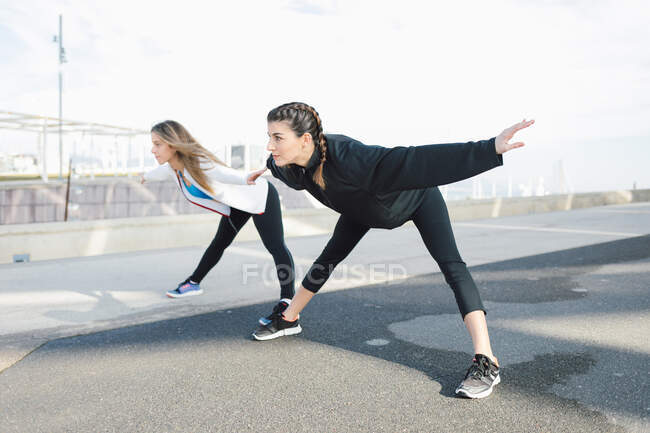 Full body fit young females in activewear bending forward on road while warming up before intense working out — Fotografia de Stock