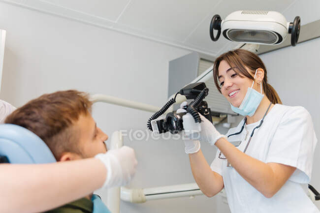 Lächelnde kompetente Zahnärztin, die Fotos männlicher Patientenzähne macht, während sie in einer modern ausgestatteten Zahnklinik arbeitet — Stockfoto