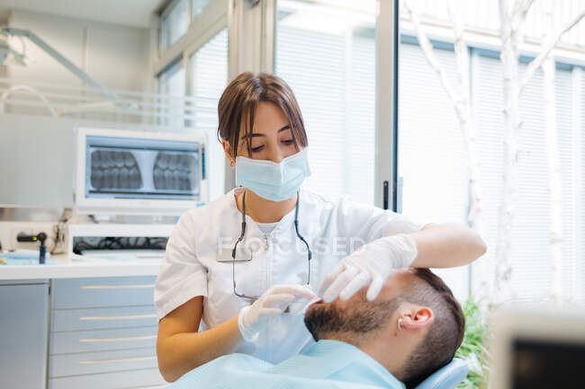 Концентрована професійна жінка-стоматолог у формі та масці, яка перевіряє стан зубів пацієнтів чоловічої статі під час роботи в сучасній стоматологічній клініці — стокове фото