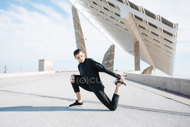 Full length determined young sportswoman in activewear stretching legs while warming up on road in suburb — Stock Photo