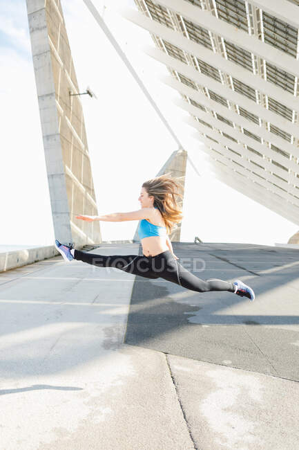 Full body flexible fit sportswoman in activewear performing split in air while working out near creative concrete construction in sunny suburb — Foto stock