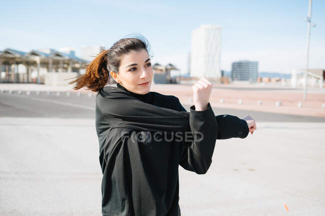 Positive fit young female in black sportswear stretching arms while warming up on sunny road in suburb and looking away contentedly - foto de stock