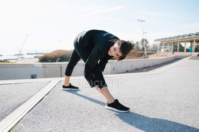 Full length determined young sportswoman in activewear stretching legs while warming up on road in suburb - foto de stock