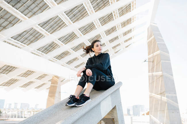 Мечтательная женщина во всю длину подходит в активной одежде, отдыхает под креативной бетонной конструкцией и смотрит в солнечный день — стоковое фото