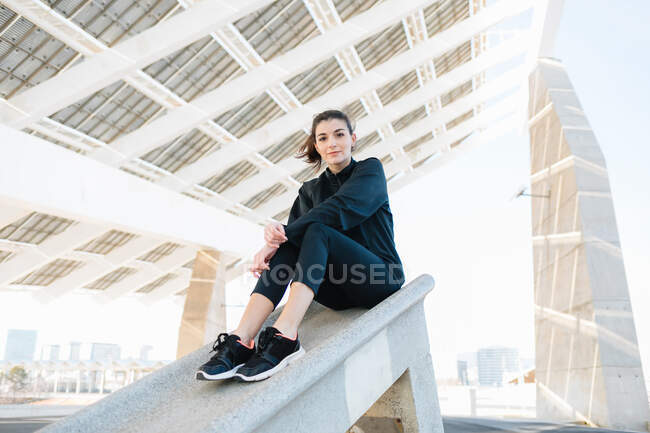 Full length dreamy fit female in activewear resting under creative concrete construction and looking at camera on sunny day — Stock Photo
