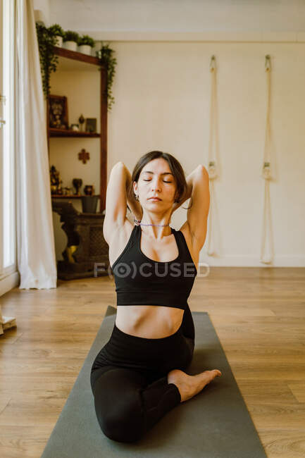 Side view of young barefoot female in sportswear sitting in Eka Pada Rajakapotasana pose while practicing yoga on mat in house - foto de stock