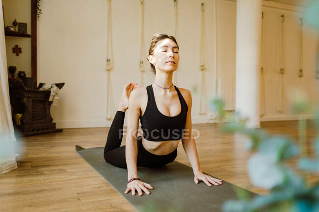 Молодая сконцентрированная женщина в спортивной одежде демонстрирует позу раджа бхуджангасаны с закрытыми глазами на коврике для йоги в доме — стоковое фото