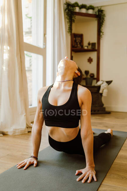 Unrecognizable flexible female in sportswear showing Bhujangasana pose while practicing yoga in house room in daylight - foto de stock