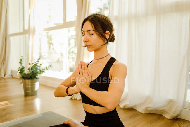 Молодая гибкая мечтательная женщина в спортивной одежде, исполняющая позу падмасаны с закрытыми глазами на коврике для йоги в доме — стоковое фото