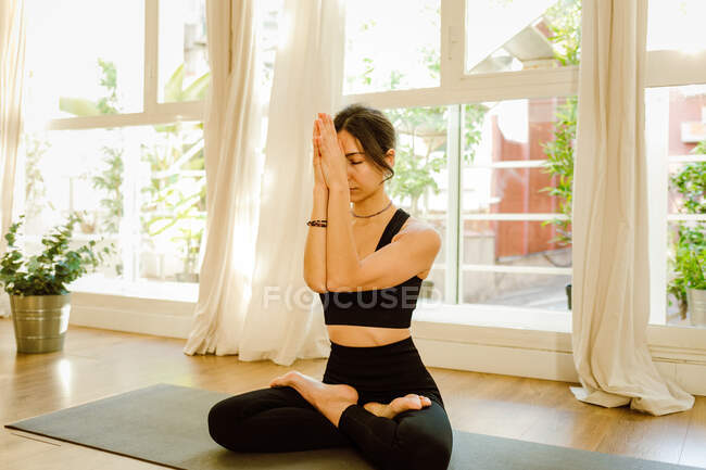 Young flexible dreamy female in sportswear performing Padmasana pose with closed eyes on yoga mat in house - foto de stock