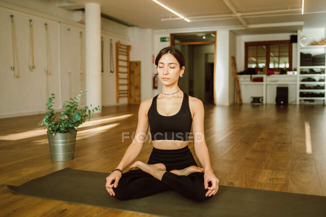 Молодая гибкая мечтательная женщина в спортивной одежде, исполняющая позу падмасаны с закрытыми глазами на коврике для йоги в доме — стоковое фото