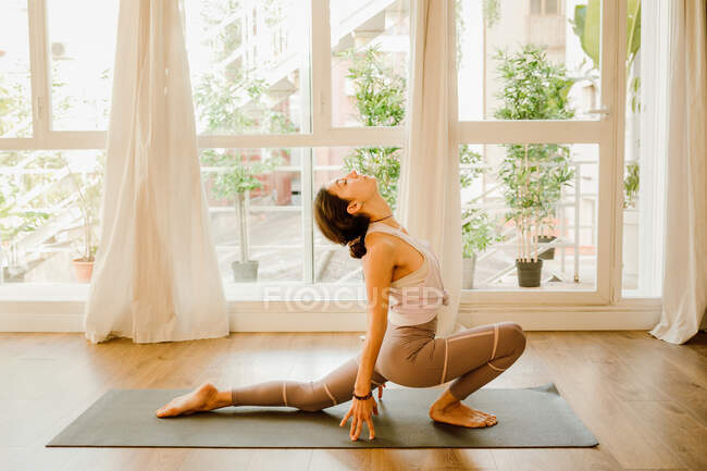 Side view of young flexible female in sportswear standing on knee and touching foot while practicing yoga in house - foto de stock