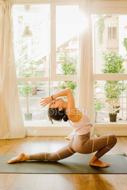 Side view of young flexible female in sportswear standing on knee and touching foot while practicing yoga in house - foto de stock