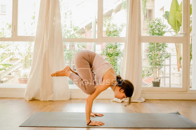 Seitenansicht einer jungen Frau in Sportbekleidung, die in Kakasana-Pose steht, während sie zu Hause Yoga gegen Fenster praktiziert — Stockfoto