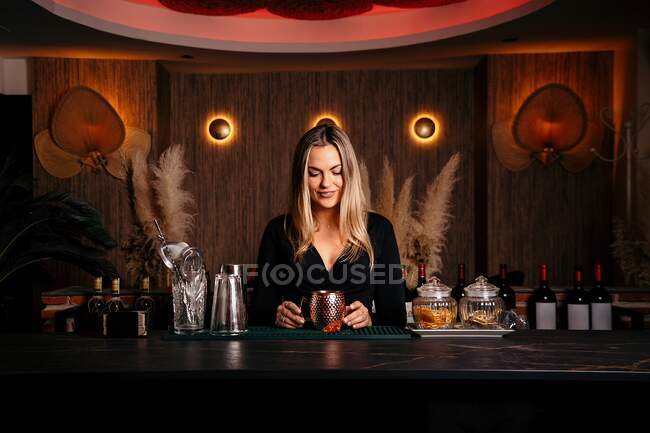 Self assured focused young female barkeeper with long blond hair in stylish outfit preparing cocktail in metal mug standing at counted in stylish bar — Foto stock