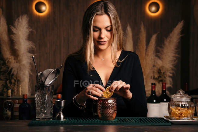 Self assured focused young female barkeeper with long blond hair in stylish outfit decorated cocktail with lemon slices while standing at counted in stylish bar — Fotografia de Stock