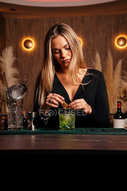 Self assured focused young female barkeeper with long blond hair in stylish outfit decorated cocktail with lemon slices while standing at counted in stylish bar — Stock Photo