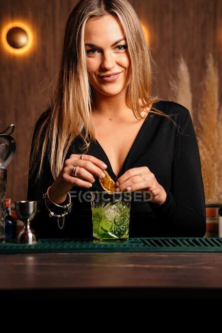 Jeune femme barman rassurée avec de longs cheveux blonds dans une tenue élégante cocktail décoré avec des tranches de citron tout en se tenant debout au compté dans le bar élégant — Photo de stock