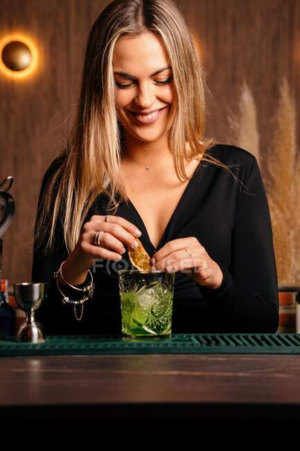 Self assured happy focused young female barkeeper with long blond hair in stylish outfit decorated cocktail with lemon slices while standing at counted in stylish bar — Fotografia de Stock