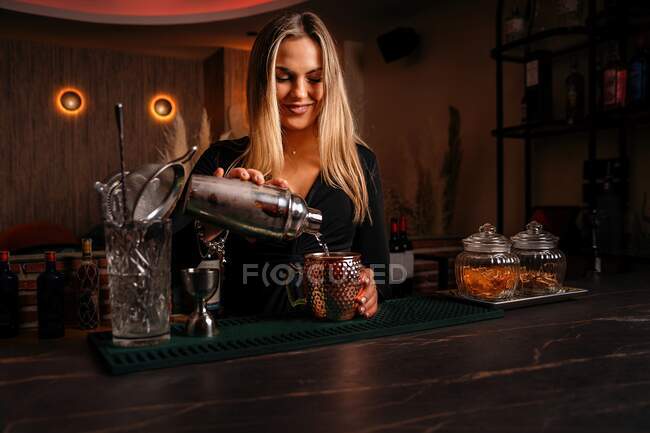 Beautiful blonde young female bartender in stylish outfit pouring alcohol cocktail from shaker into elegant metal mug in restaurant — Stock Photo