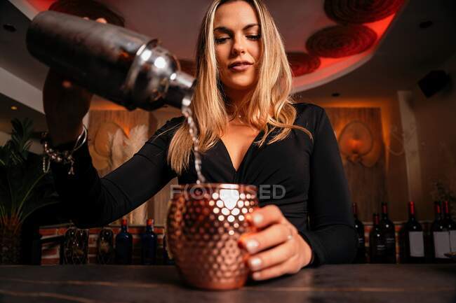 Beautiful blonde young female bartender in stylish outfit pouring alcohol  cocktail from shaker into elegant metal mug in restaurant — mix, sensual -  Stock Photo | #458253834