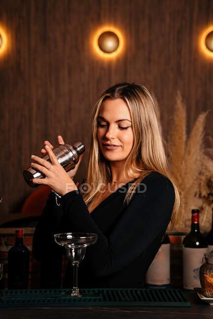 Belle jeune femme barman avec de longs cheveux blonds dans des vêtements élégants souriant tout en mélangeant cocktail dans shaker dans le bar moderne — Photo de stock