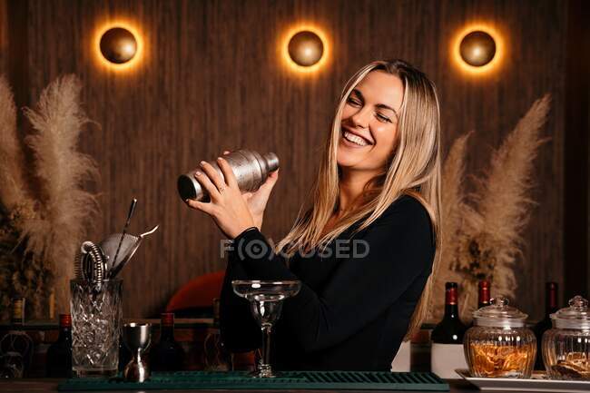 Joyeux jeune femme barman avec de longs cheveux blonds dans des vêtements élégants souriant tout en mélangeant cocktail dans shaker dans le bar moderne — Photo de stock