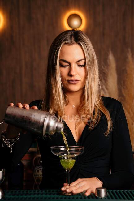 Bella bionda giovane barista in abito elegante versando cocktail alcolici da shaker in elegante vetro margarita nel ristorante — Foto stock