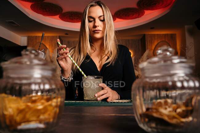 Blonde attractive female bartender stirring iced alcohol cocktail with long straw serving on wooden counter — Stock Photo