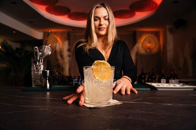 Содержание молодой барменши с длинными светлыми волосами, подающей холодный алкогольный коктейль, украшенный лимонным ломтиком в стильном ресторане — стоковое фото