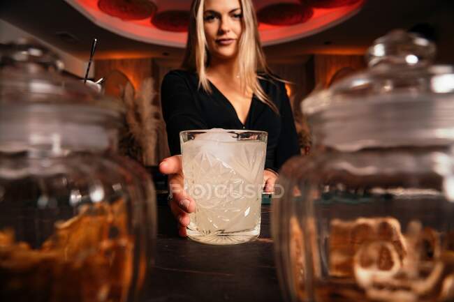 Content young female bartender with long blond hair serving cold alcohol cocktail in stylish restaurant looking at camera — Stock Photo