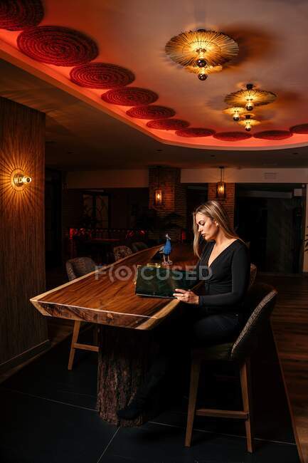 Vista lateral de un joven cliente femenino concentrado en un elegante atuendo sentado en una mesa de madera en un elegante bar y menú de lectura - foto de stock