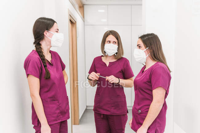 Grupo de jovens enfermeiros de conteúdo em uniforme médico e máscaras conversando no corredor hospitalar moderno — Fotografia de Stock