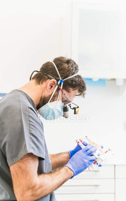 Visão lateral focada técnico odontológico masculino em uniforme médico e óculos de lupa binocular trabalhando com próteses dentárias na clínica moderna — Fotografia de Stock
