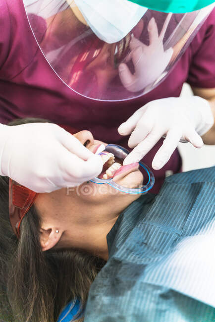 Crop unrecognizable dentist in gloves and face shield examining female patient teeth in light contemporary dental clinic — Stock Photo