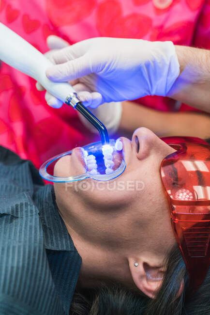 Recadrer un dentiste anonyme dans des gants à l'aide d'un outil de polymérisation aux ultraviolets dentaires pendant l'intervention avec un patient en clinique — Photo de stock