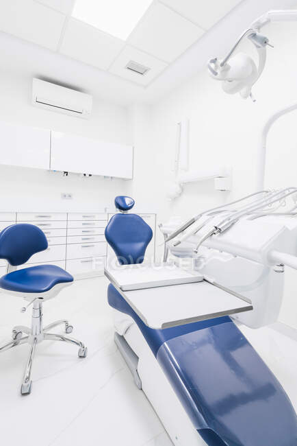 Интерьер современной аккуратной стоматологической клиники с синим стулом и белой мебелью, оборудованной современной стоматологической машиной и инструментами — стоковое фото