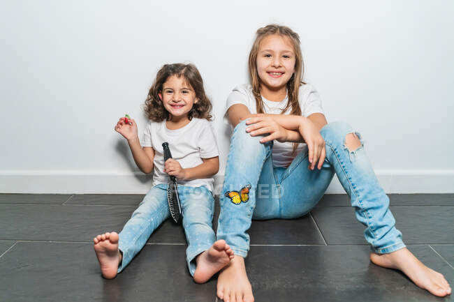 Счастливые милые маленькие сестры в белой рубашке и джинсах сидят на полу у стены дома и смотрят в камеру с зубастыми улыбками — стоковое фото