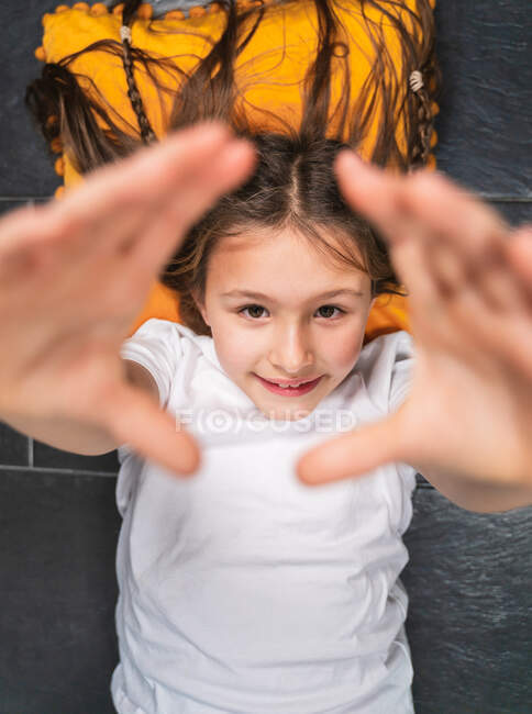 De cima positivo bonito menina em camisa branca deitada no travesseiro no chão e levantando as mãos para a câmera — Fotografia de Stock