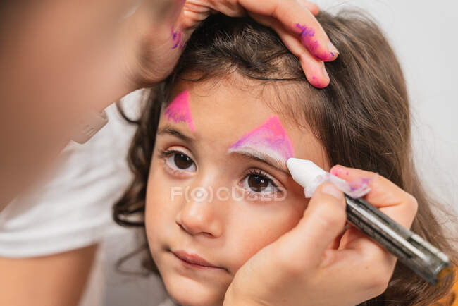 Crop artista creativo aplicando coloridas pinturas de arte corporal en la cara linda niña en el estudio de luz - foto de stock