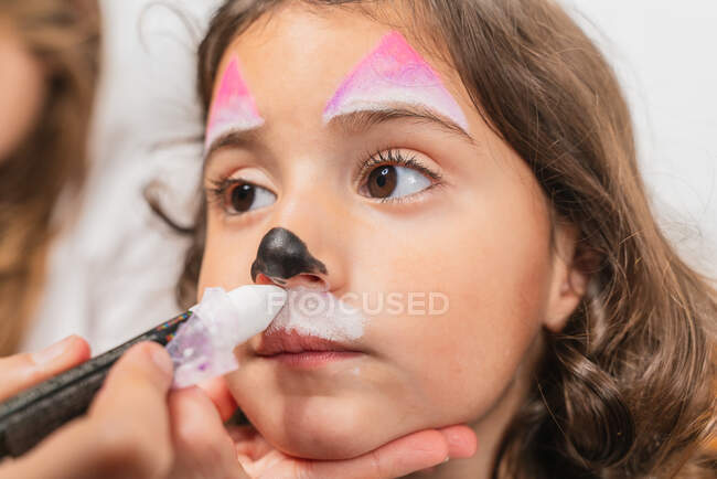 Crop creative artist applying colorful body art paints on cute little girl face in light studio — Stock Photo
