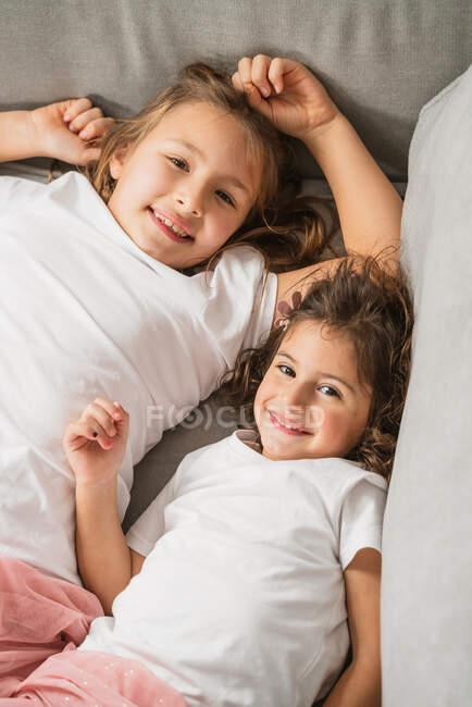 Зверху веселі маленькі сестри лежать на зручному дивані і дивляться на камеру — стокове фото