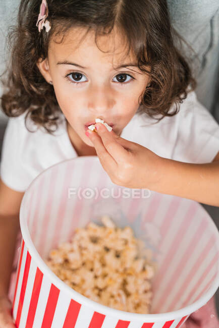 De cima adorável menina amigável em camisa branca desfrutando doce pipoca fresca e olhando para a câmera — Fotografia de Stock