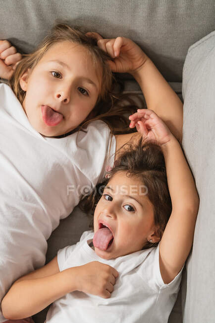 Зверху веселі маленькі сестри посміхаються і показують мови, лежачи на зручному дивані і дивлячись на камеру — стокове фото